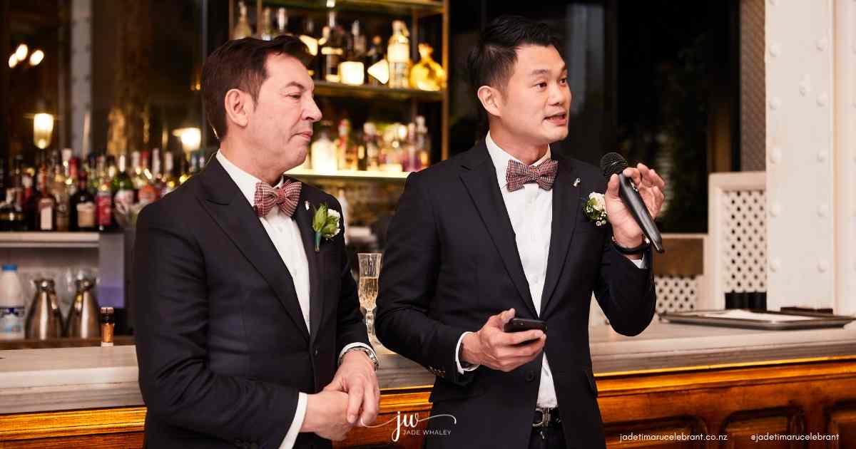 Two men who are grooms, dressed in suits with tartan bowties, standing up at their wedding, leaning against a bar. The Asian man is speaking into a microphone, holding his phone. Timaru Wedding Celebrant Jade Whaley, NZ.