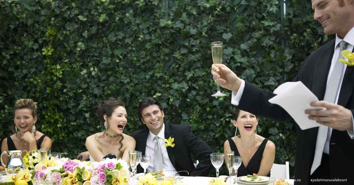 The wedding reception table has yellow flowers, plates and wine glasses. A man in a black and white suit is speaking, talking into a microphone and holding white paper. The bride, groom and two bridesmaids wearing black dresses are laughing. Timaru Wedding Celebrant Jade Whaley, NZ.