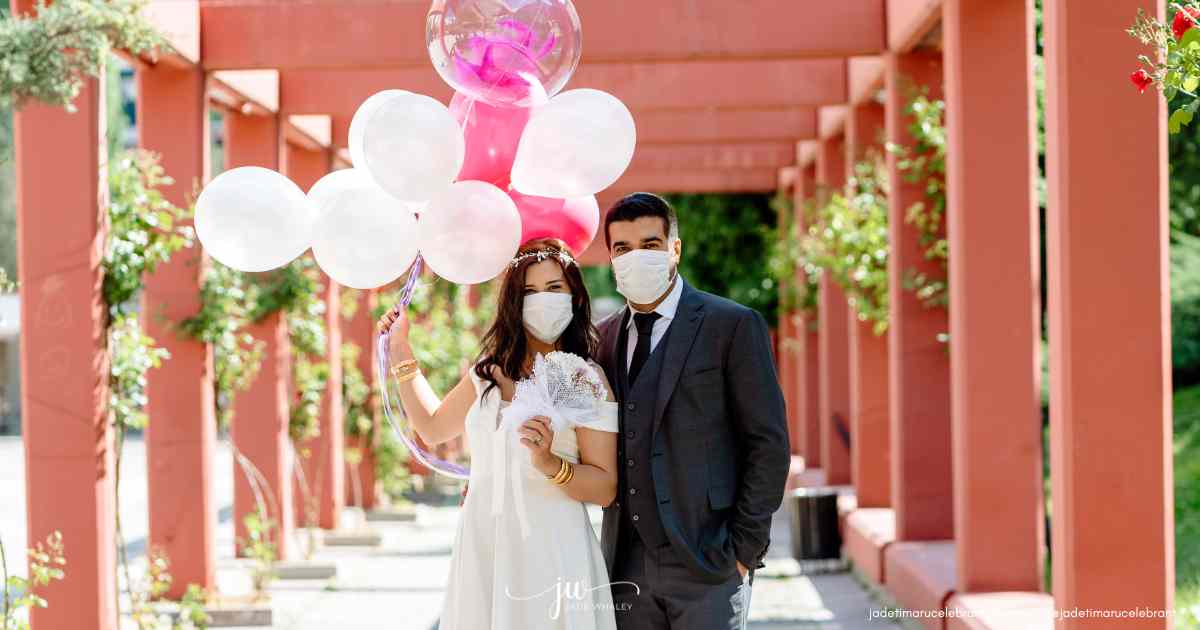 A bride and Groom are standing under a pink wooden gazebo, each wearing a protective facemask to stop COVID-19 spread and holding pink balloons. Timaru Wedding Celebrant Jade Whaley, NZ.