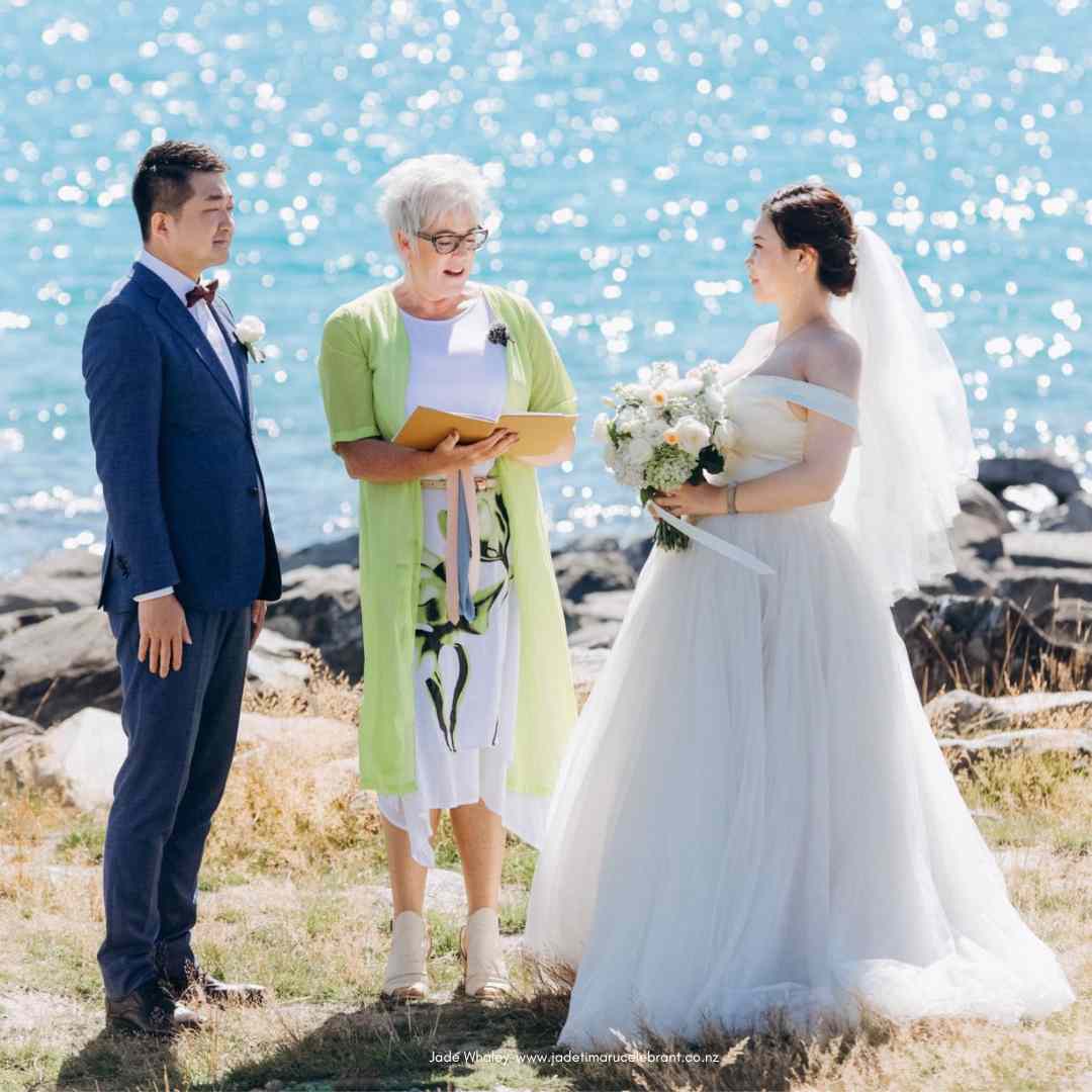 Elopement at Lake Tekapo with Jade Whaley Celebrant. Bride and groom standing on shores of Lake Tekapo with wedding celebrant reading out of gold folder.